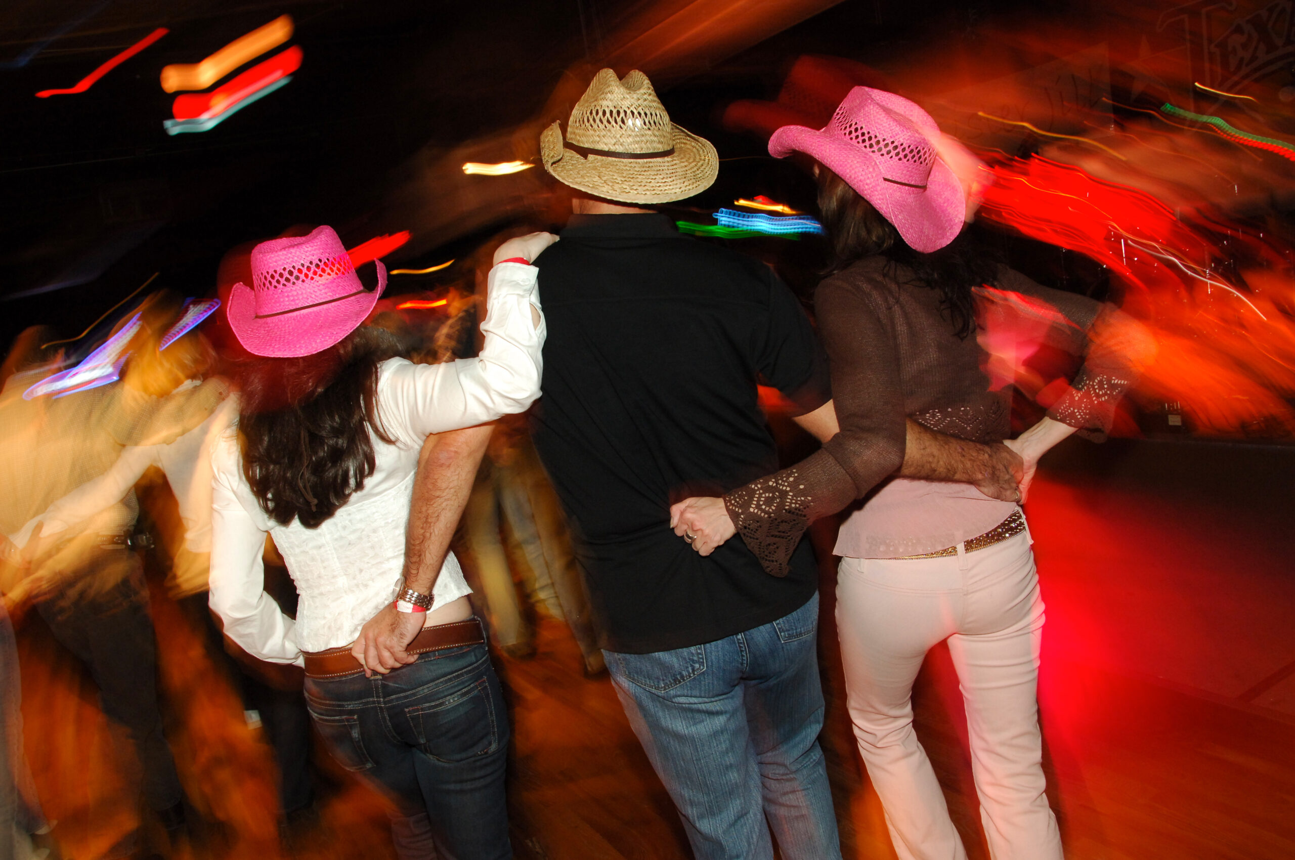 Three people line dancing, two women wearing pink cowboy hats and a man in the middle wearing a straw hat, with their arms around each other in a lively, colorful setting.