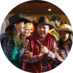 Group of smiling people in cowboy hats enjoying a line dance, with a man in a red vest and brown hat at the center, surrounded by friends in a warm, cozy setting.